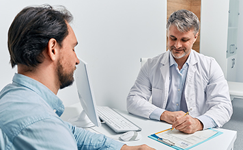 Dos personas sentadas junto a un escritorio en una consulta médica. Uno, el médico, lleva una bata blanca. En el escritorio hay documentos y equipo médico.